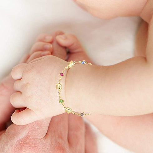 A baby's hand wearing the Good Luck Elephant Bracelet with Multicolor Birthstones gently rests on an adult's hand. In the background, the baby's toes are visible, adding to the heartwarming scene.